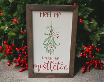 May include: A rectangular wooden framed sign with the text "Meet Me Under The Mistletoe" in red script. The sign features a green mistletoe illustration with red berries on a white background. The frame is a dark brown color.