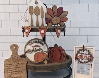 May include: A tiered tray with wooden fall-themed decorations. Includes a tag with utensils, a turkey, a banner, a cutting board with a recipe, a round sign with pumpkins, a pumpkin, and a door with a wreath that says "gather".