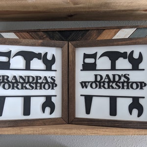 May include: Two framed signs with a white background and dark brown frames. Each sign features black silhouettes of tools and the words "GRANDPA'S WORKSHOP" and "DAD'S WORKSHOP". The signs are displayed on a wooden shelf.