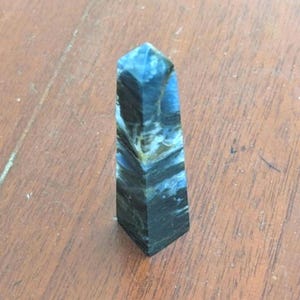 May include: A blue and black tiger's eye obelisk crystal point. The crystal has a pointed top and a flat base.