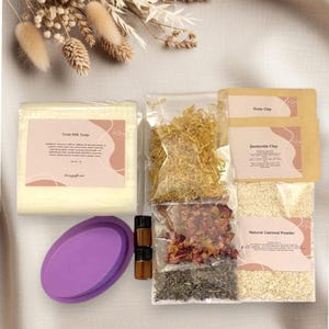 May include: A collection of natural skincare products, including a bar of Goat Milk Soap, dried flowers, rose clay, bentonite clay, natural oatmeal powder, essential oils, and a purple oval mold. The products are arranged on a neutral-toned surface.
