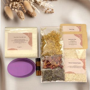May include: A collection of natural skincare ingredients, including a bar of shea butter goat milk soap, dried flowers, rose clay, bentonite clay, natural oatmeal powder, and essential oil bottles. A purple oval soap mold is also included.