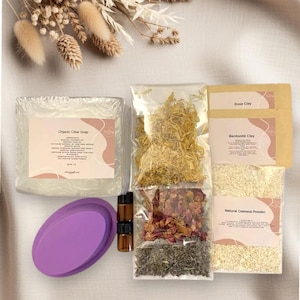 Puede incluir: Un surtido de productos naturales para el cuidado de la piel, que incluye una pastilla de jabón transparente, flores secas, arcilla de rosa y bentonita, polvo de avena natural y botellas de aceites esenciales. También se incluye un jabonera ovalada morada.