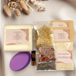 Könnte beinhalten: Ein Hautpflege-Set mit einem Sheabutter-Seifenstück, einer lila Seifenschale, getrockneten Blumen, Rosenlehm, Bentonit-Lehm, natürlichem Haferflockenpulver und kleinen bernsteinfarbenen Flaschen. Auf neutralem Untergrund angeordnet.