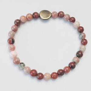 May include: A beaded bracelet featuring round, polished beads in shades of pink, red, and cream. The bracelet is strung together with a metal clasp. The beads have a natural, speckled appearance, and the bracelet is displayed on a white surface.