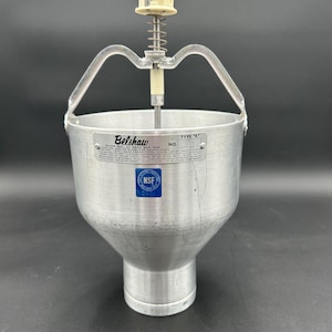 May include: A silver metal donut batter dispenser with a spring-loaded handle and a white plastic top. The dispenser has a label that reads "Belshaw TYPE "K"" and "NSF".