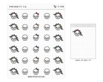 May include: A sticker sheet from Paperbits Co. labeled ST-265M featuring rows of small skull illustrations, some with floral crowns and bows. A size reference box indicates a width of 1.5 inches for the individual skull sticker design.