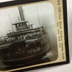 以下が含まれることがあります： 人々でいっぱいのフェリーの写真。右側に「44-5286-New Jersey ferryboat bringing crowds into New York City.」というテキストがあります。