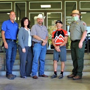 Puede incluir: Cinco personas están de pie en un tramo de escaleras fuera de un edificio. La persona de la derecha lleva un uniforme verde y un sombrero de guardabosques. La persona del medio lleva un maillot de ciclismo rojo y negro. Las otras tres personas llevan ropa informal.
