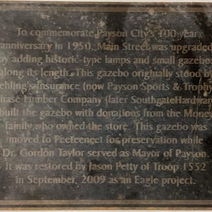 May include: A black metal plaque with a silver inscription commemorating the 100th anniversary of Payson City in 1950. The plaque describes the history of a gazebo built by Chase Lumber Company and donated by the Money family. It was moved to Peeteneet for preservation and restored by Jason Petty in 2009.