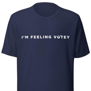 May include: A navy blue short-sleeved t-shirt featuring the white text "I'M FEELING VOTEY" printed across the chest. The letter O in the word VOTEY is replaced by a white checkmark symbol. The shirt has a crew neckline and a standard fit.