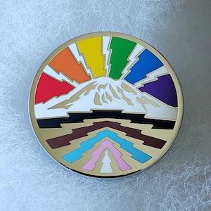 May include: A round, gold-colored pin with a white mountain peak surrounded by a rainbow with lightning bolts. The bottom of the pin features a brown, black, blue, and pink geometric design.