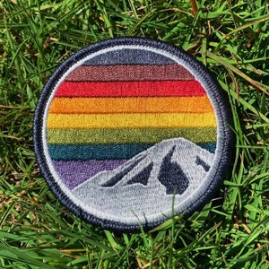 May include: A round embroidered patch with a rainbow mountain design. The patch features a rainbow with seven colors, a white mountain peak, and a black outline.