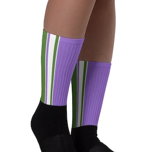 Puede incluir: Un par de calcetines a rayas moradas y verdes con puños negros en los tobillos. Los calcetines se llevan puestos en los pies de una persona.