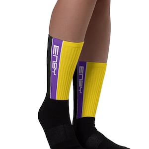 Puede incluir: Un par de calcetines negros y amarillos con una franja morada y la palabra "Embly" escrita en el lateral.