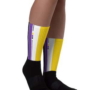 Puede incluir: Un par de calcetines negros con rayas amarillas y moradas. Los calcetines tienen un fondo blanco con una fina línea negra que separa las rayas.