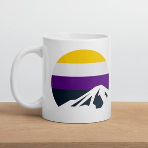 Puede incluir: Taza de cerámica blanca con un diseño circular amarillo, blanco, morado y negro. El diseño circular representa una montaña estilizada con un patrón de bandera.