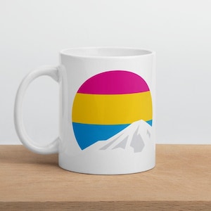 May include: White ceramic mug with a graphic of a mountain and a circle with the colors of the pansexual pride flag: pink, yellow, and blue.