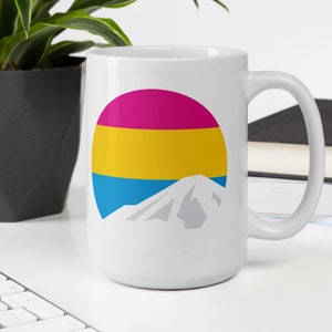May include: White ceramic mug with a graphic of a mountain and a circle with the colors of the pansexual pride flag: pink, yellow, and blue.