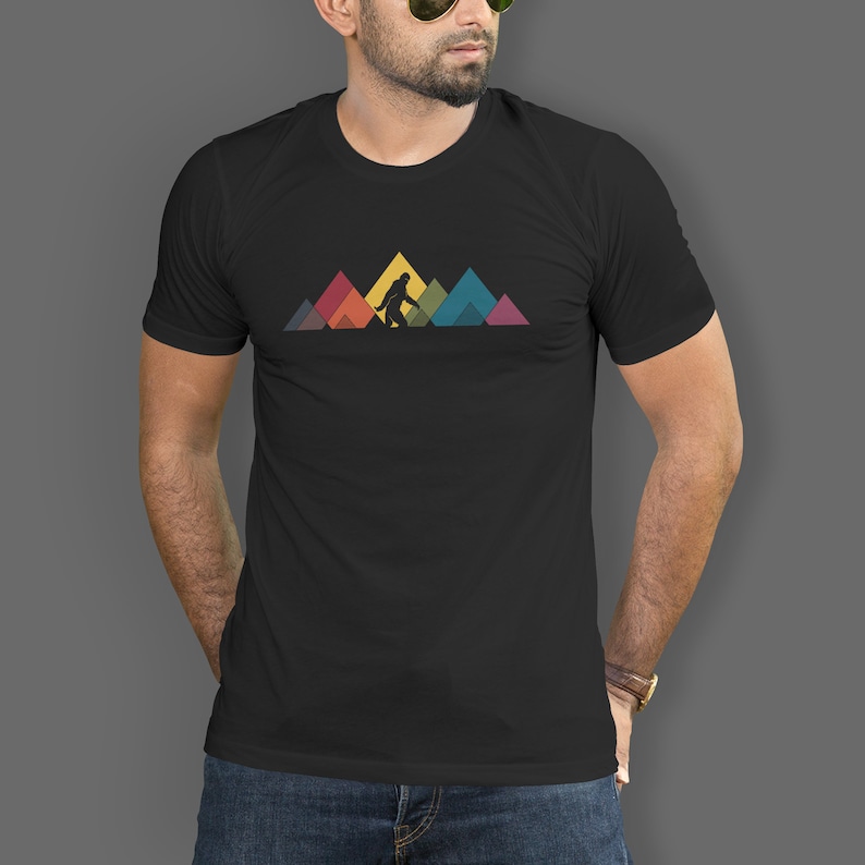 May include: A black t-shirt featuring a graphic of Bigfoot walking in front of a row of colourful mountains.
