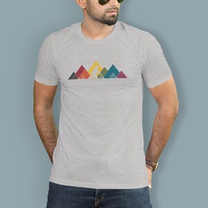 May include: A heather grey t-shirt featuring a graphic of a stylised Bigfoot walking across a mountain range. The mountains are made up of colourful geometric shapes.