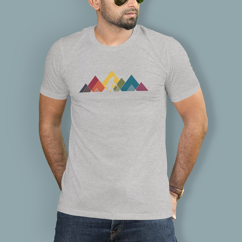 May include: A heather grey t-shirt featuring a graphic of a stylised Bigfoot walking across a mountain range. The mountains are made up of colourful geometric shapes.