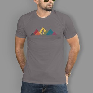May include: A grey t-shirt featuring a graphic of Bigfoot walking in front of a mountain range. The mountains are made up of colourful geometric shapes.