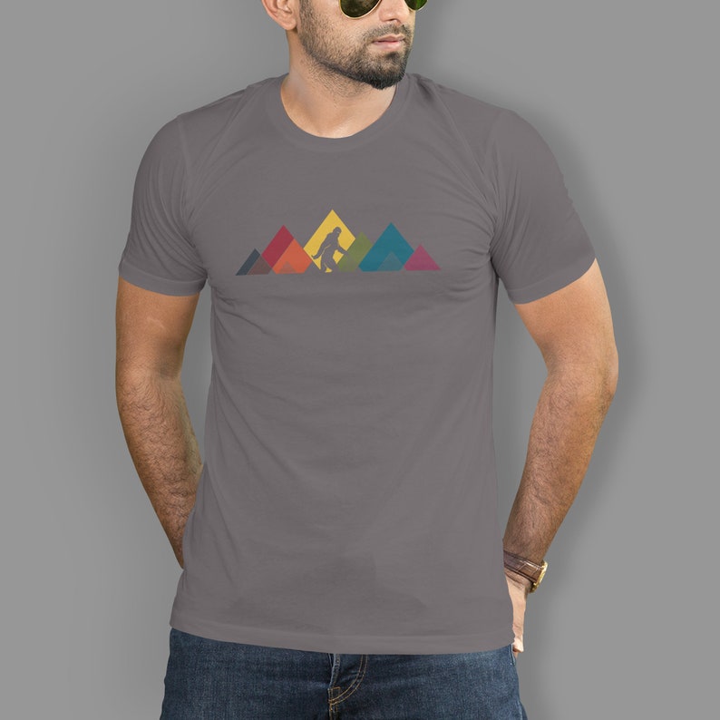 May include: A grey t-shirt featuring a graphic of Bigfoot walking in front of a mountain range. The mountains are made up of colourful geometric shapes.