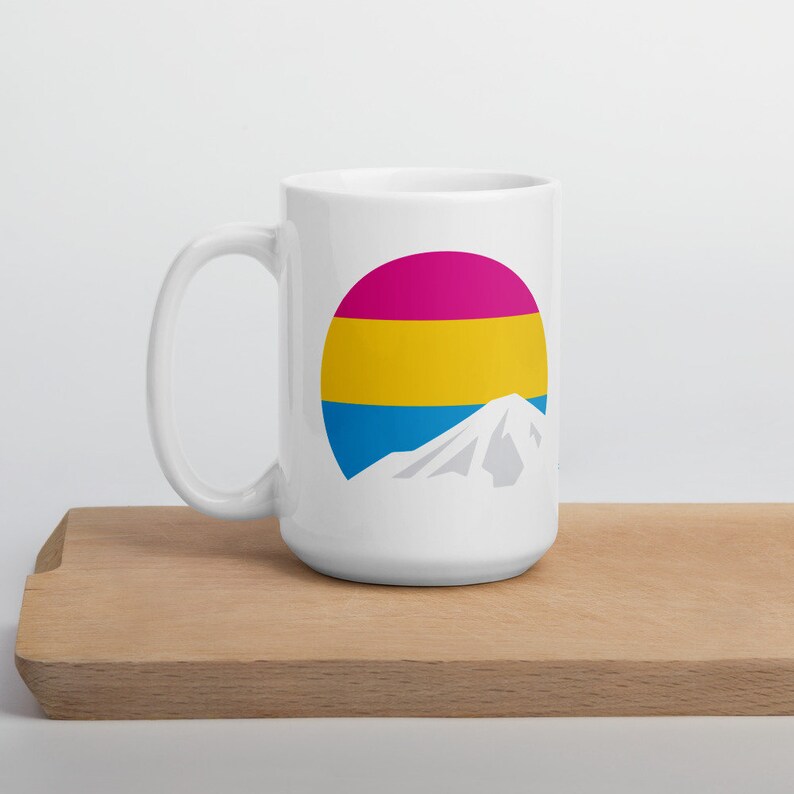 May include: White ceramic mug with a graphic of a mountain peak and a circle with the colors of the pansexual pride flag: pink, yellow, and blue.