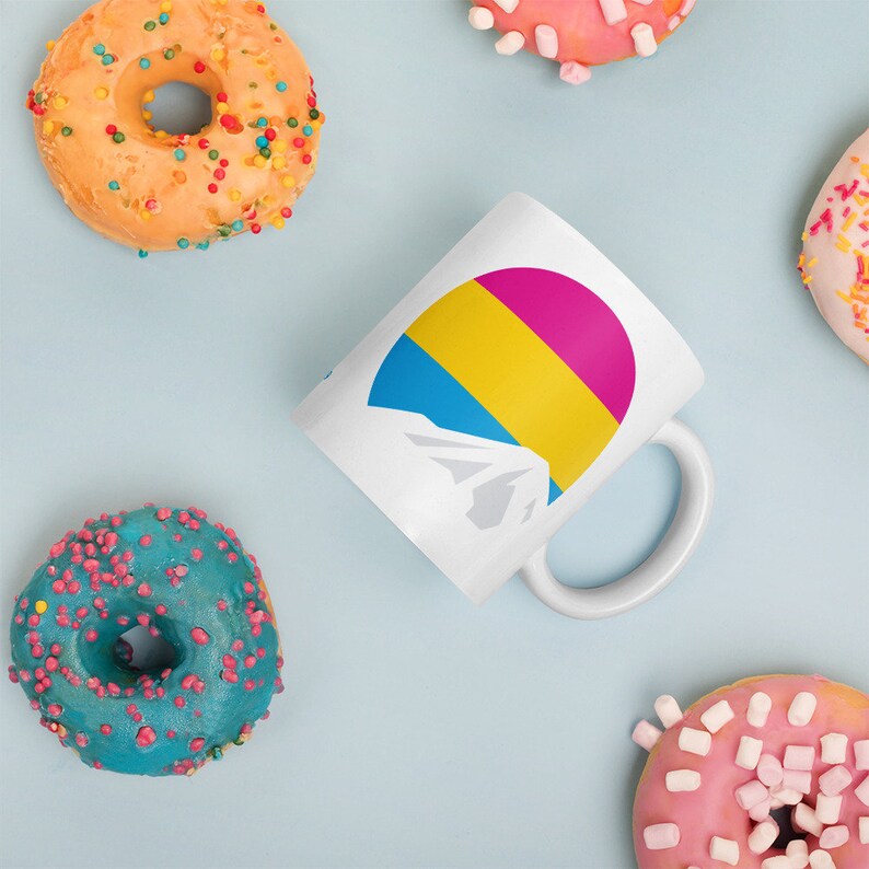 May include: White ceramic mug with a pansexual pride flag design. The flag is a half circle with blue, yellow, and pink stripes. The mug is surrounded by donuts with colorful sprinkles and marshmallows.
