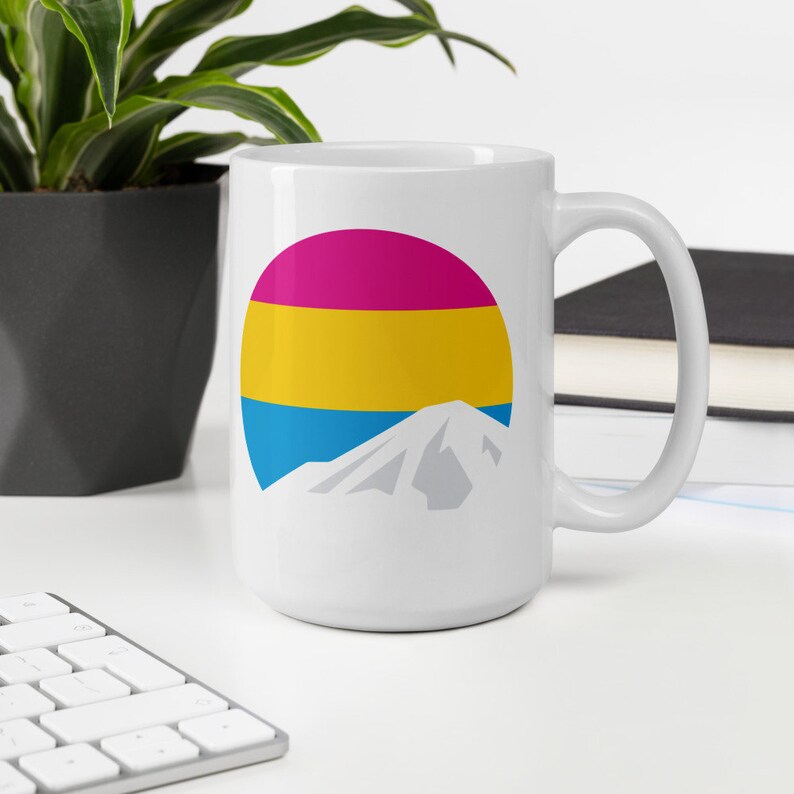 May include: White ceramic mug with a graphic of a mountain and a circle with the colors of the pansexual pride flag: pink, yellow, and blue.