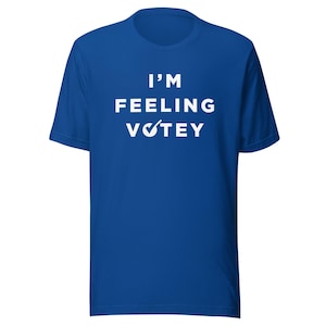 Puede incluir: Una camiseta de manga corta de color azul real con texto blanco centrado en el pecho que dice I'M FEELING VOTEY, con un símbolo de marca de verificación blanca que reemplaza la letra O en la palabra VOTEY. Tiene cuello redondo y corte estándar.