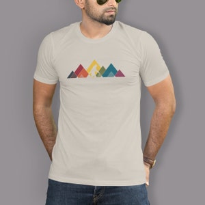 May include: A beige t-shirt featuring a graphic of Bigfoot walking across a mountain range. The mountains are made up of colourful geometric shapes.