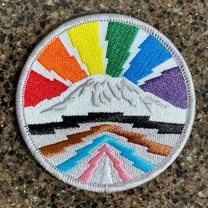 Peut inclure: Un patch brodé blanc avec un motif de montagne arc-en-ciel. La montagne est entourée d'un arc-en-ciel avec des couleurs rouge, orange, jaune, vert, bleu, indigo et violet. Le bas du patch présente un motif géométrique brun, bleu et rose.