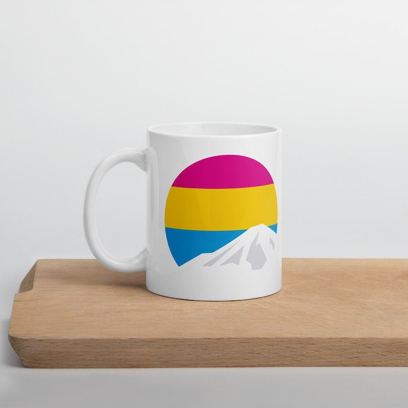 May include: White ceramic mug with a graphic of a mountain and a circle with the colors of the pansexual pride flag: pink, yellow, and blue.