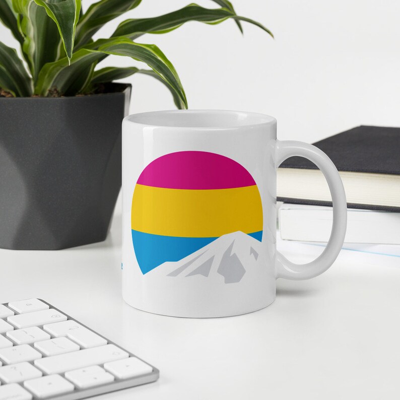 May include: White ceramic mug with a graphic of a mountain and a circle with horizontal stripes of pink, yellow, and blue.