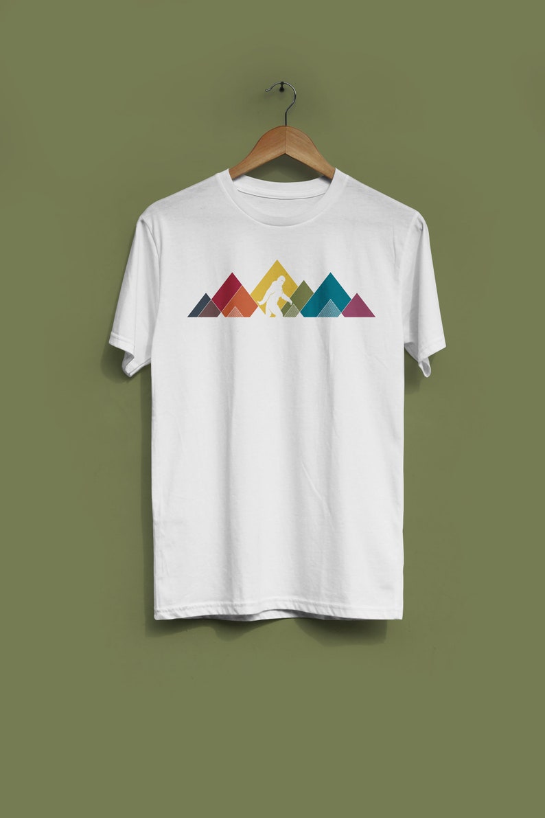 May include: White t-shirt featuring a graphic of a Bigfoot walking in front of a mountain range. The mountains are made up of colourful geometric shapes.