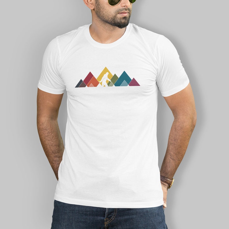 May include: White t-shirt featuring a graphic of a Bigfoot walking across a mountain range. The mountains are made up of colourful geometric shapes.