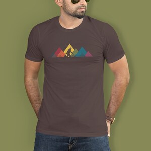 May include: A brown t-shirt featuring a graphic of Bigfoot walking across a mountain range. The mountains are made up of colourful triangles.