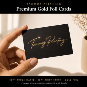 Puede incluir: Una tarjeta de visita rectangular negra con el texto Tammoz Printing en una fuente de escritura de l&aacute;mina de oro met&aacute;lico. La tarjeta tiene un acabado mate y se sostiene entre los dedos, mostrando la calidad reflectante de las letras doradas sobre el fondo oscuro.