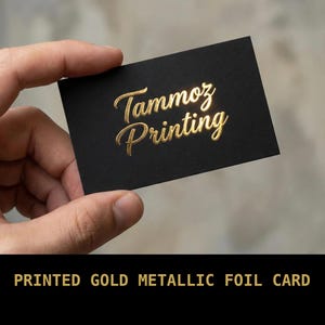 Puede incluir: Una tarjeta de visita rectangular negra con el texto Tammoz Printing en una fuente de escritura de l&aacute;mina de oro met&aacute;lico. La tarjeta tiene un acabado mate y las letras doradas est&aacute;n en relieve, creando una superficie reflectante.