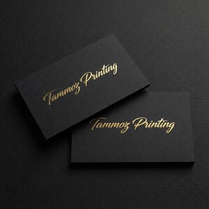 Puede incluir: Dos tarjetas de visita rectangulares de cartulina negra mate. Cada tarjeta presenta el texto Tammoz Printing en relieve con una fuente cursiva dorada met&aacute;lica. Las tarjetas est&aacute;n colocadas en &aacute;ngulo, con una tarjeta superpuesta parcialmente a la otra.