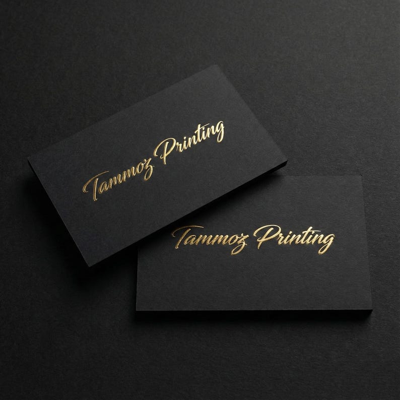Puede incluir: Dos tarjetas de visita rectangulares de cartulina negra mate. Cada tarjeta presenta el texto Tammoz Printing en relieve con una fuente cursiva dorada met&aacute;lica. Las tarjetas est&aacute;n colocadas en &aacute;ngulo, con una tarjeta superpuesta parcialmente a la otra.