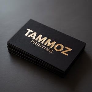 Puede incluir: Una pila de tarjetas de visita rectangulares con un acabado negro mate. La tarjeta superior presenta el texto TAMMOZ PRINTING grabado con un acabado de l&aacute;mina de oro met&aacute;lico. Las letras son negritas y est&aacute;n centradas en la cartulina negra.