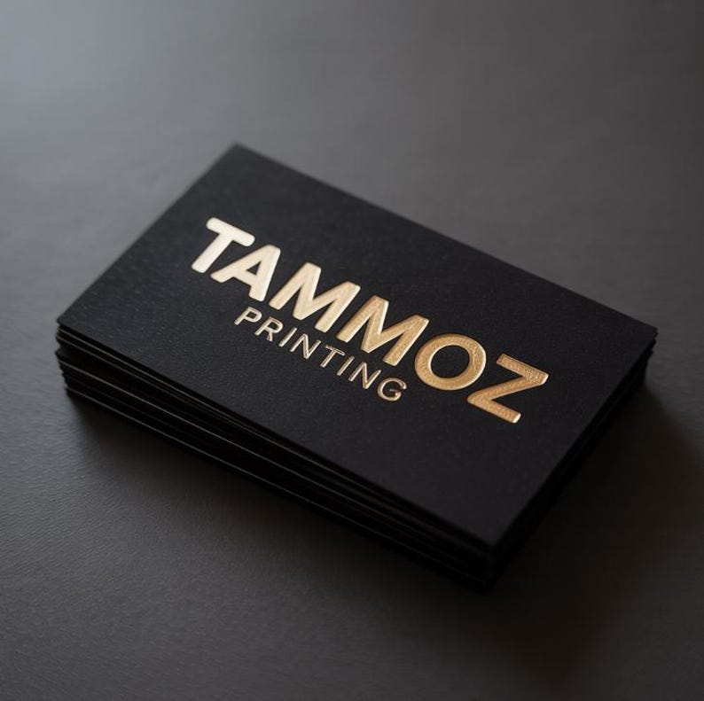 Puede incluir: Una pila de tarjetas de visita rectangulares con un acabado negro mate. La tarjeta superior presenta el texto TAMMOZ PRINTING grabado con un acabado de l&aacute;mina de oro met&aacute;lico. Las letras son negritas y est&aacute;n centradas en la cartulina negra.