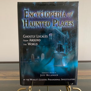 May include: A blue book titled "Encyclopedia of Haunted Places: Ghostly Locales From Around The World" compiled and edited by Jeff Belanger. The book cover features a blurry image of a house with a graveyard in the foreground.