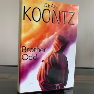 May include: A book cover with a dark purple and orange background. The title "Brother Odd" is written in black text. The author's name "Dean Koontz" is written in silver text. A person wearing a dark purple hooded robe is standing in front of a cloudy sky.