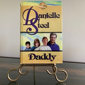 May include: A paperback book titled "Daddy" by Danielle Steel. The book cover features a photo of a family with a yellow background and blue sky. The book is on a gold metal stand.