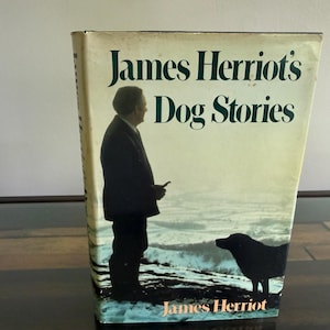 May include: A hardcover book titled James Herriot's Dog Stories with a dust jacket featuring a silhouette of a person standing next to a dog in a snowy landscape. The cover text is printed in dark green and white against a pale background.