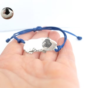 Può includere: Un braccialetto in corda blu con un ciondolo d'argento raffigurante un porcellino d'India bianco e nero.